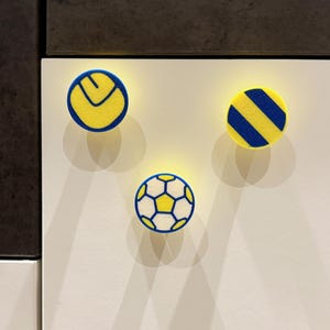 May include: Three circular magnets with sports designs. One has a yellow circle with a blue curved line, another has blue diagonal stripes on yellow, and the third is a football design with white, yellow, and blue.