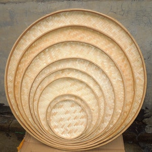 May include: A collection of six round, woven bamboo trays of varying sizes, nested together. The trays have a natural, light brown color and a textured surface. The largest tray is approximately 20 inches in diameter.