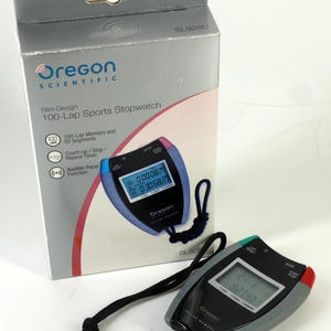 Op de afbeelding: Een zwarte en grijze Oregon Scientific 100-Lap Sports Stopwatch met een digitale display die de tijd weergeeft. De stopwatch heeft blauwe, rode en groene accenten en een zwart koord. Op de doos staat "Slim Design 100-Lap Sports Stopwatch".