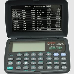 Peut inclure: Calculatrice noire Aurora TM100B avec un couvercle ouvert affichant un tableau de conversion métrique. La calculatrice possède de nombreux boutons et un écran numérique. Elle fait également office de réveil.