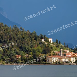 Può includere: Una vista pittoresca di un piccolo villaggio incastonato su una collina che domina un lago. Alberi verdi e lussureggianti e montagne circondano il villaggio, creando un'ambientazione serena e idilliaca.