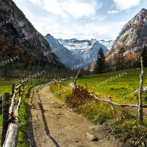 Può includere: Un sentiero sterrato si snoda attraverso una valle verde con una recinzione in legno sul lato sinistro. Il sentiero conduce a una catena montuosa in lontananza con cime innevate. Le montagne sono circondate da alberi con foglie autunnali.