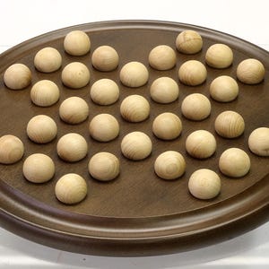Pode incluir: Um jogo de solitário de madeira com um tabuleiro oval marrom escuro. O tabuleiro tem uma superfície de jogo rebaixada e é preenchido com 33 bolas de madeira. As bolas são de cor clara e esféricas.