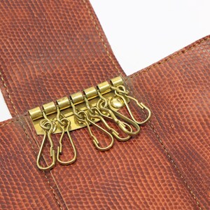 May include: A brown leather key holder with a textured surface and gold-colored key hooks. The key holder is open, revealing the key hooks and the interior lining. The stitching is visible, adding to the design.