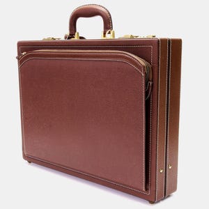 Pode incluir: Uma pasta de couro castanho com uma pega e ferragens douradas. A pasta tem um bolso frontal com zíper e costuras brancas. As bordas também são costuradas em branco.