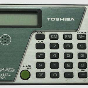 May include: A vintage Toshiba LC-854WA liquid crystal calculator. The calculator has a green and silver color scheme with a digital display, a keypad with black buttons, and the Toshiba logo. It features alarm and set functions.