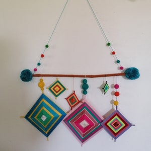 Peut inclure: Une décoration murale colorée avec des œuvres d'art en forme de losange en fil de différentes tailles et couleurs, notamment le bleu, le rose et le vert. La pièce est suspendue à une cheville en bois avec des pompons turquoise et un chapelet de perles colorées.