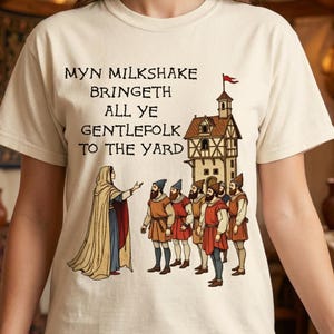 以下が含まれることがあります： クリーム色のTシャツ。中世風のイラストとテキストが特徴です。イラストは、ローブを着た人物が男性のグループと建物を示しています。テキストは「MYN MILKSHAKE BRINGETH ALL YE GENTLEFOLK TO THE YARD」と書かれています。