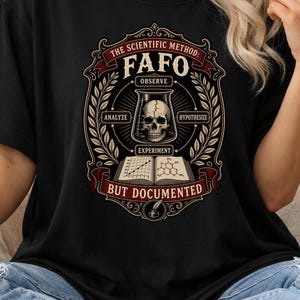May include: A black t-shirt featuring a graphic design with the text "THE SCIENTIFIC METHOD: FAFO BUT DOCUMENTED". The design includes a skull inside a beaker, an open book with scientific diagrams, and decorative laurel leaf borders.