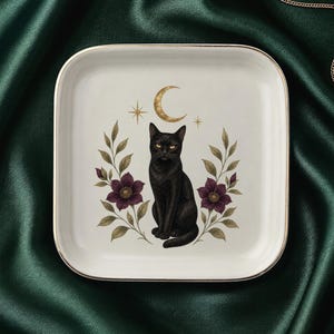 May include: A square white ceramic tray with a gold-toned rim featuring a central illustration of a black cat sitting upright. The cat is flanked by two purple flowers on leafy stems, with a gold crescent moon and two small stars positioned above.