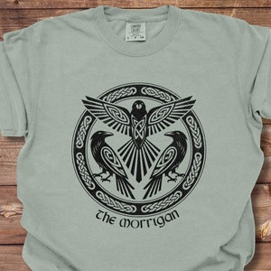 May include: A light green t-shirt with a black and white graphic of a Celtic knot design with a bird and two ravens. The text "the morrigan" is printed below the design.
