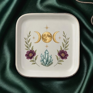 May include: A square white ceramic trinket dish with a gold-toned rim. The center features a graphic illustration of a golden triple moon phase symbol, a cluster of light blue crystals, and two floral stems with dark purple flowers and green leaves.