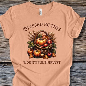 Puede incluir: Camiseta color melocotón con un diseño de cosecha de otoño. El gráfico incluye una cesta llena de calabazas, manzanas y trigo, con las palabras "Blessed Be This Bountiful Harvest" en una fuente decorativa.