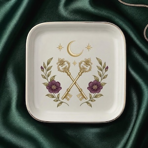 May include: A square white ceramic trinket dish with a gold-toned rim. The center features a graphic illustration of two crossed ornate gold keys, a crescent moon, small stars, and two symmetrical floral branches with dark purple flowers.