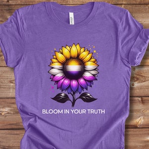 Puede incluir: Camiseta morada con un girasol amarillo con un centro morado, blanco y amarillo. El texto "Bloom in your truth" está impreso debajo del girasol.