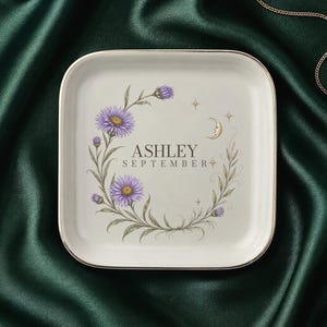 May include: A square white ceramic trinket dish with a gold-toned rim. The center features a floral wreath of purple aster flowers, a crescent moon, and small stars. The text ASHLEY and SEPTEMBER is printed in the center of the floral design.