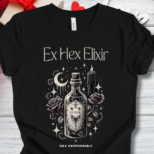 Puede incluir: Camiseta negra con un gráfico detallado de una botella de poción etiquetada "Elixir", con un diseño de mariposa, rosas, una luna creciente y estrellas. También se muestra el texto "Ex Hex Elixir" y "Hex Responsibly".