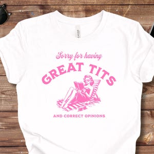 May include: White t-shirt with a pink graphic that reads "Sorry for having GREAT TITS and CORRECT OPINIONS." The graphic features a woman reclining and reading. The shirt is on a wooden surface.