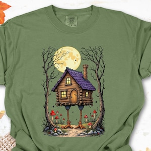 May include: Olive green t-shirt featuring a whimsical illustration of a house on stilts, with a purple roof, set against a full moon. The design includes trees, mushrooms, and a path, creating a fantasy theme.