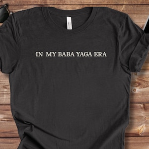 May include: Black t-shirt with the white text "IN MY BABA YAGA ERA". The shirt is displayed on a wooden surface with sunglasses, a smartphone, and sneakers in the background.