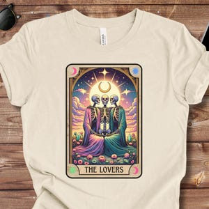 May include: Cream-colored t-shirt featuring a tarot card design. The card depicts three skeletons in colorful robes, with the text "THE LOVERS" at the bottom. The design includes a celestial background with a crescent moon and stars.