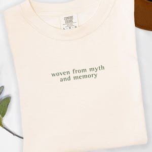 May include: Cream-coloured t-shirt with the text "woven from myth and memory" in green. The shirt is a Comfort Colors brand, with a crew neck and short sleeves. A brown hat is partially visible in the background.