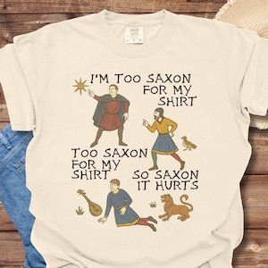 以下が含まれることがあります： 中世風のイラストと「I'm too Saxon for my shirt. Too Saxon for my shirt. So Saxon it hurts.」というテキストが描かれた白いTシャツです。