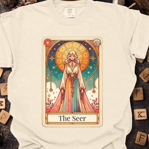 May include: Cream-colored t-shirt featuring a tarot card design. The card depicts a woman in a colorful gown with a celestial background and the text "The Seer". The shirt is displayed on a wooden surface with scattered runes.