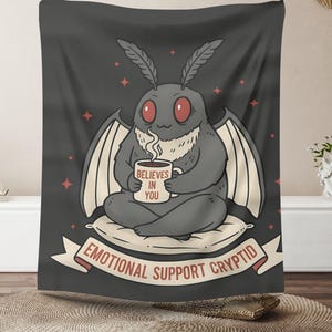 Könnte beinhalten: Dunkelgraue Tapisserie mit einer Cartoon-Mottenkreatur mit roten Augen, die eine Tasse mit der Aufschrift "Believes in You" hält. Die Motte sitzt auf einem Kissen, darunter steht "Emotional Support Cryptid" auf einem Banner.