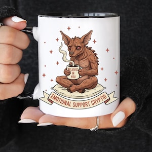 Puede incluir: Taza de cerámica blanca con interior y asa negros, con un criptido de dibujos animados sosteniendo una taza de café que dice "Believes in You". La taza está decorada con estrellas y las palabras "Emotional Support Cryptid".