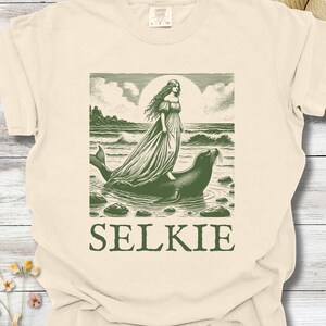 May include: Cream-colored t-shirt featuring a green illustration of a woman standing on a seal in the ocean, with the word "SELKIE" below. The design includes a sun, waves, and rocks, creating a mythical theme.