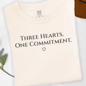 Op de afbeelding: Crèmekleurig T-shirt met de zwarte tekst "THREE HEARTS. ONE COMMITMENT." en een klein hartsymbool. Het shirt is opgevouwen en heeft een label met "COMFORT COLORS". Op de achtergrond is een bruine hoed te zien.