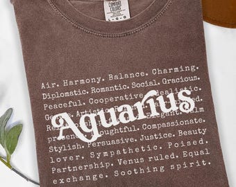Könnte beinhalten: Ein braunes Comfort Colors T-Shirt mit dem weißen Wort "Aquarius" und beschreibenden Wörtern. Im Hintergrund ist ein brauner Hut zu sehen. Das T-Shirt ist gefaltet und hat die Größe L-G-G. Das Shirt ist aus weichem Material.