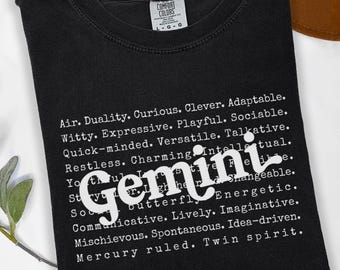 Könnte beinhalten: Schwarzes T-Shirt mit dem weißen Wort "Gemini", umgeben von einer Liste von Eigenschaften. Das Shirt ist gefaltet und hat ein Comfort Colors-Etikett. Ein brauner Hut ist im Hintergrund.