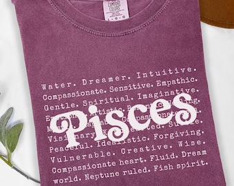 Könnte beinhalten: Burgunderfarbenes Comfort Colors T-Shirt mit dem Wort "Pisces" in weißer Schrift. Das Shirt zeigt eine Liste von Eigenschaften des Sternzeichens Fische in weißem Text, darunter "Träumer", "Intuitiv" und "Kreativ".