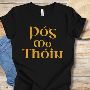 May include: Black t-shirt with the text "Pós mo Thóin" in gold lettering.