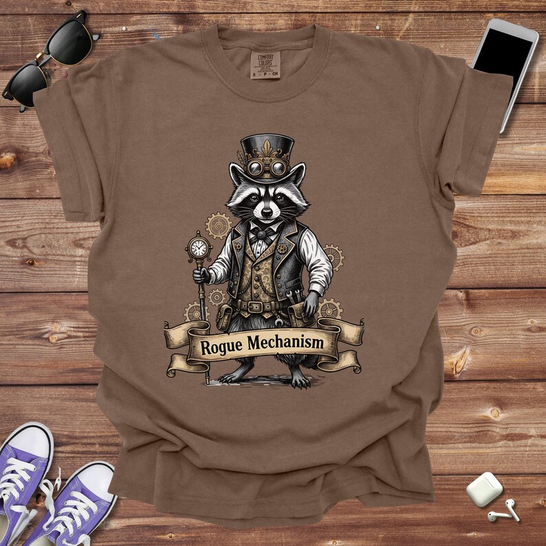 May include: Brown t-shirt featuring a raccoon in a steampunk outfit, including a top hat, goggles, and a vest. The raccoon holds a pocket watch and the banner reads "Rogue Mechanism".