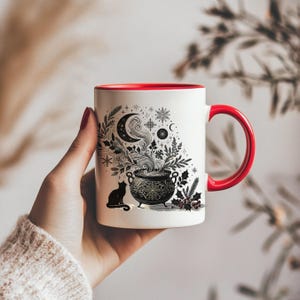 Op de afbeelding: Witte keramische mok met een rode binnenkant en oor. De mok heeft een zwart-wit illustratie van een ketel met stoom, een maan, sneeuwvlokken en een zwarte kat. Het ontwerp heeft een grillig, magisch thema.