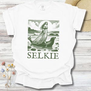 May include: White t-shirt with a green illustration of a woman in a flowing gown standing on a seal in the ocean. The word "SELKIE" is printed in green below the image. The shirt is made of a soft material.