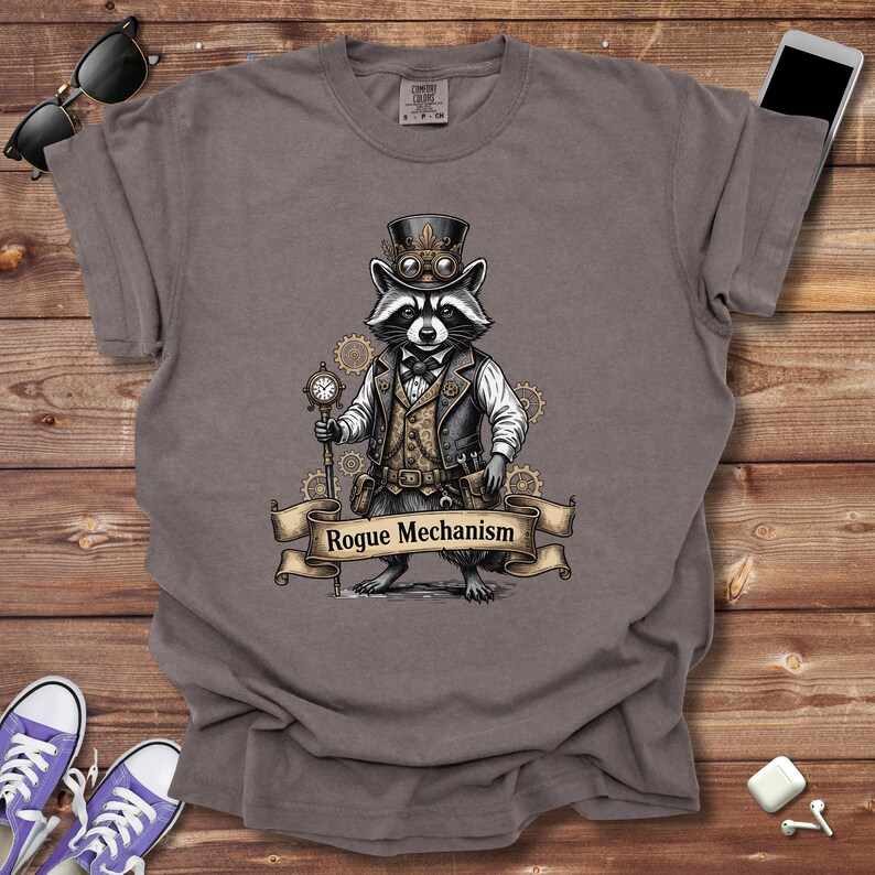 May include: A gray t-shirt featuring a raccoon dressed in steampunk attire, including a top hat and vest, holding a pocket watch. The text "Rogue Mechanism" is displayed on a banner below the raccoon.