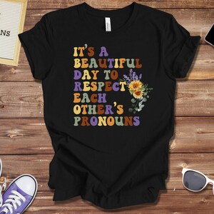 May include: Black t-shirt with the text "It's a beautiful day to respect each other's pronouns" in rainbow colors. The text is surrounded by flowers and leaves.