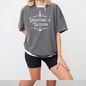 Puede incluir: Una camiseta gris oscuro de gran tamaño con un gráfico blanco que dice "Unsociable & Taciturn" en una fuente decorativa.