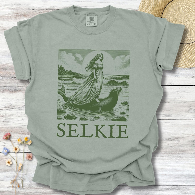 May include: Sage green t-shirt featuring a graphic of a woman riding a seal in the ocean, with the word "SELKIE" below. The design is in shades of green, evoking a vintage aesthetic. The shirt is made of a soft, comfortable material.