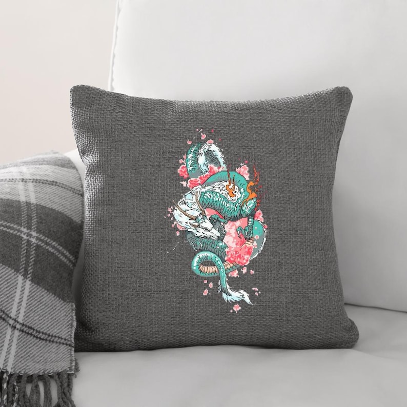 May include: Grey cushion featuring a detailed illustration of a teal and white dragon intertwined with pink cherry blossoms. The dragon has orange accents and a long, winding body. The cushion is square and made of a textured fabric.