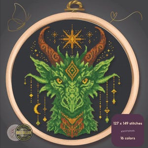 May include: Cross-stitch embroidery of a green dragon with orange horns and accents, set against a dark gray background. The design is framed in a wooden hoop. The text reads "127 x 149 stitches" and "16 colors".