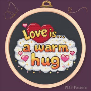 Puede incluir: Un diseño de punto de cruz en un aro de madera presenta la frase "Love is... a warm hug" en letras naranjas y amarillas, con un corazón rojo y corazones rosas. El fondo es gris oscuro.