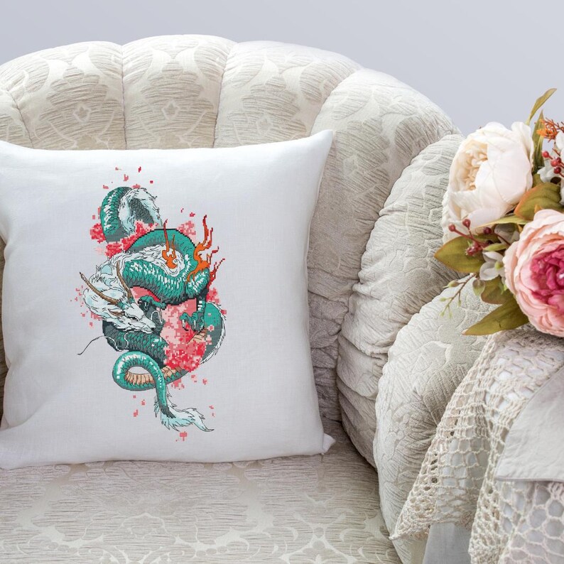 May include: A white square cushion with a detailed illustration of a teal and white dragon intertwined with pink cherry blossoms. The dragon has orange accents. The cushion is on a patterned beige armchair.
