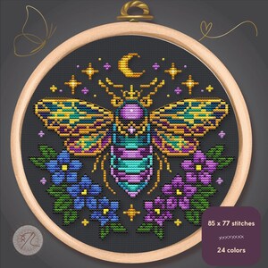 Puede incluir: Un bordado de punto de cruz en un aro de madera presenta un insecto estilizado con alas coloridas, una luna creciente y estrellas sobre un fondo gris oscuro. Flores azules y moradas flanquean el insecto. El texto indica 85 x 77 puntadas y 24 colores.