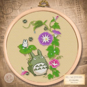 Op de afbeelding: Kruissteekborduurwerk in een houten hoepel met een groen Totoro-personage, een klein wit wezen en levendige paarse en roze windrozen. Het ontwerp bevat groene bladeren en kleine zwarte accenten op een beige achtergrond.