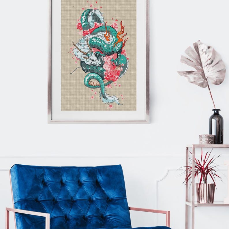 May include: Framed artwork showing a dragon illustration. The dragon is teal, white, and orange, with pink cherry blossoms. The artwork is set against a beige grid background and framed in silver. The artwork is displayed in a room with a blue chair.